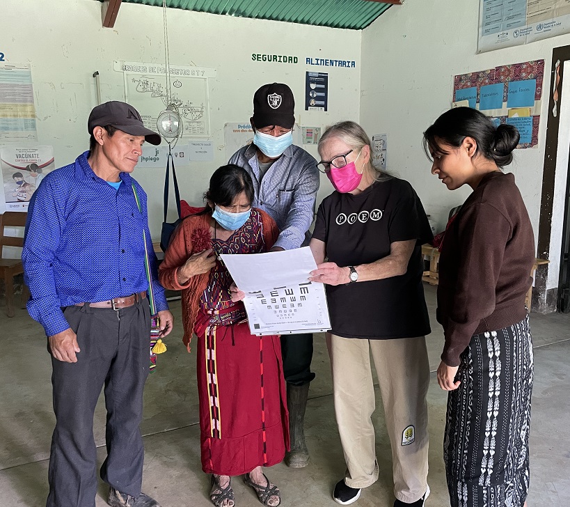 Darkskinned man in a cap and blue button-down shirt stands beside a woman wearing Ixil huipil, skirt and face mask. She and an older woman pale-skinned woman with glasses and facemask hold a poster. Man in mask and young woman look at poster.
